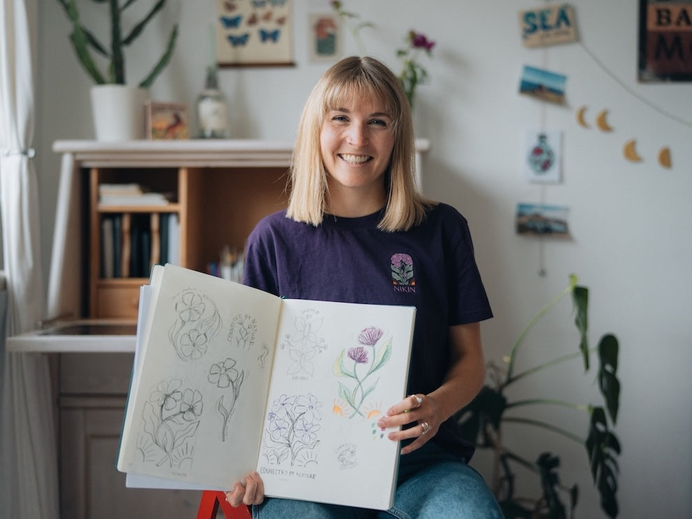 Sophia Hultsch is sitting in her studio, smiling at the camera and holding an open sketchbook with floral designs in her hand. She is wearing a purple NIKIN TreeShirt with the "Connected by Nature" design.