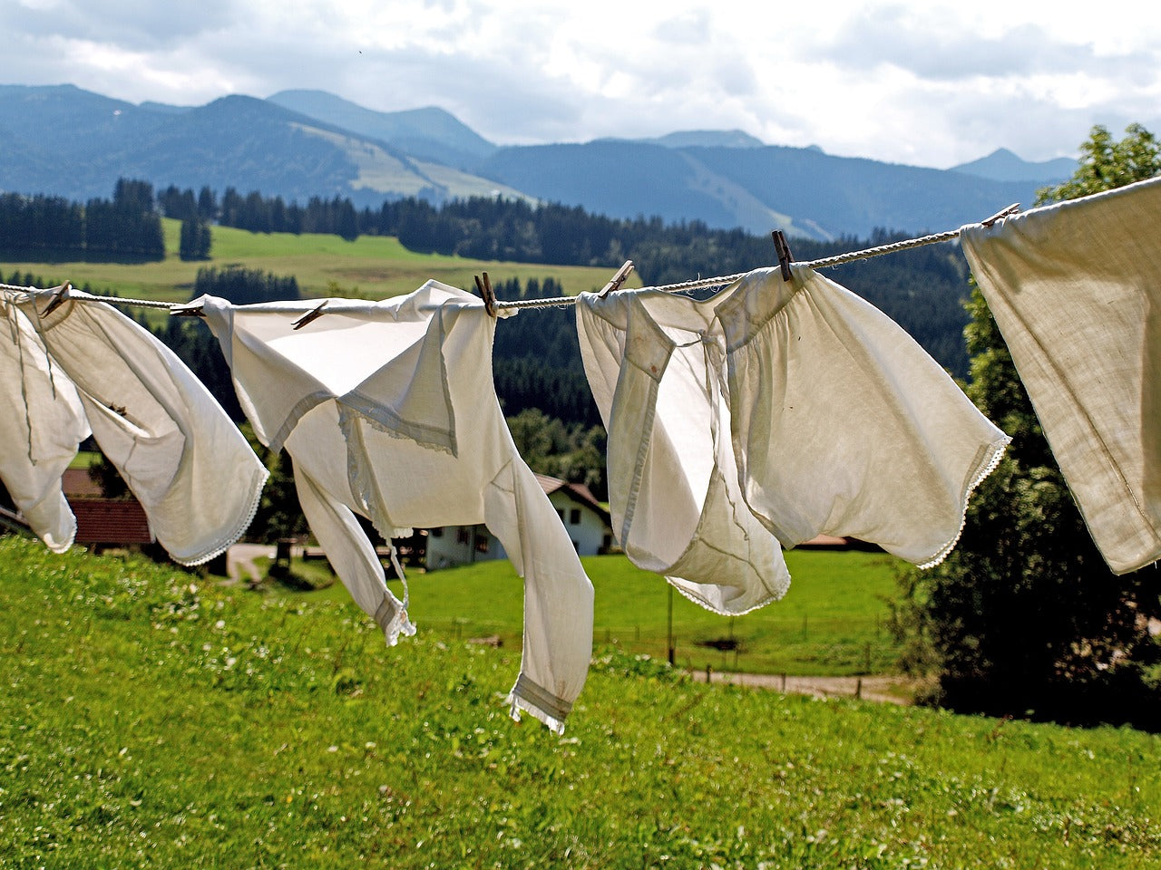 Weiße Wäsche auf einer Wäscheleine im Wind, mit grünen Wiesen, Bauernhäusern und Bergen im Hintergrund – nachhaltiges Trocknen in der Natur.